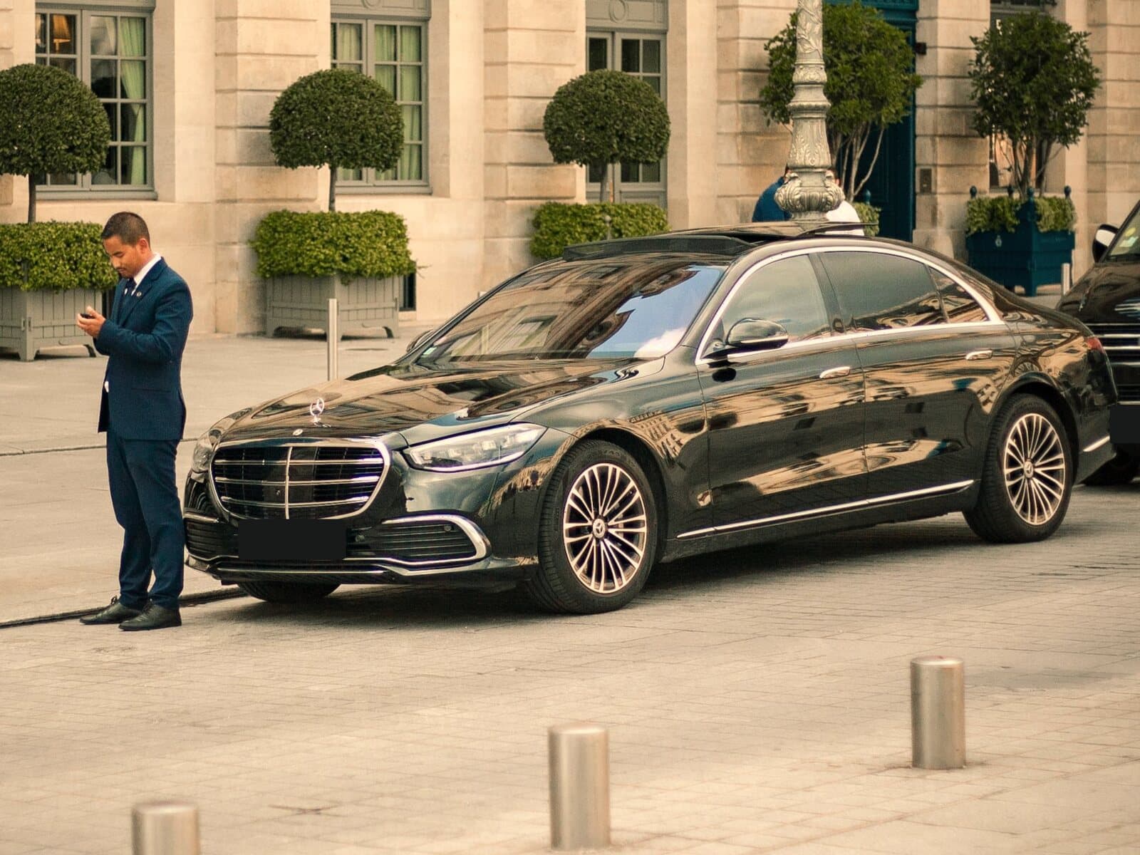 A chauffeur in a suit waits beside a black sedan outside a stone building